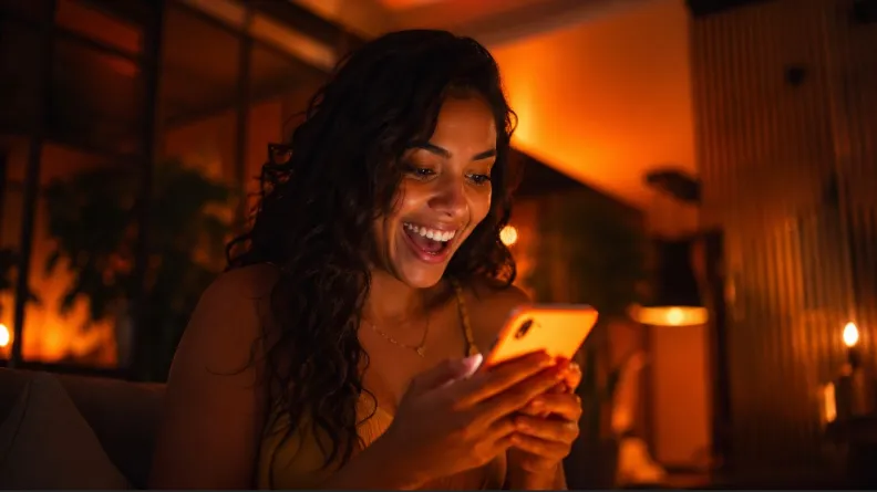 Uma mulher com expressão de surpresa e alegria olha para a tela de um smartphone em um ambiente com iluminação quente e acolhedora.
