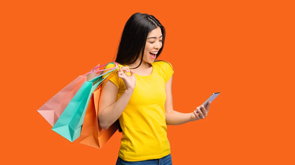 Mulher jovem com traços asiáticos e expressão alegre olha para um smartphone enquanto carrega sacolas de compras coloridas no ombro, contra um fundo laranja vibrante.