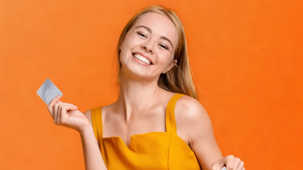 Mulher loira sorridente, vestindo uma blusa amarela, segura um cartão cinza contra um fundo laranja vibrante.