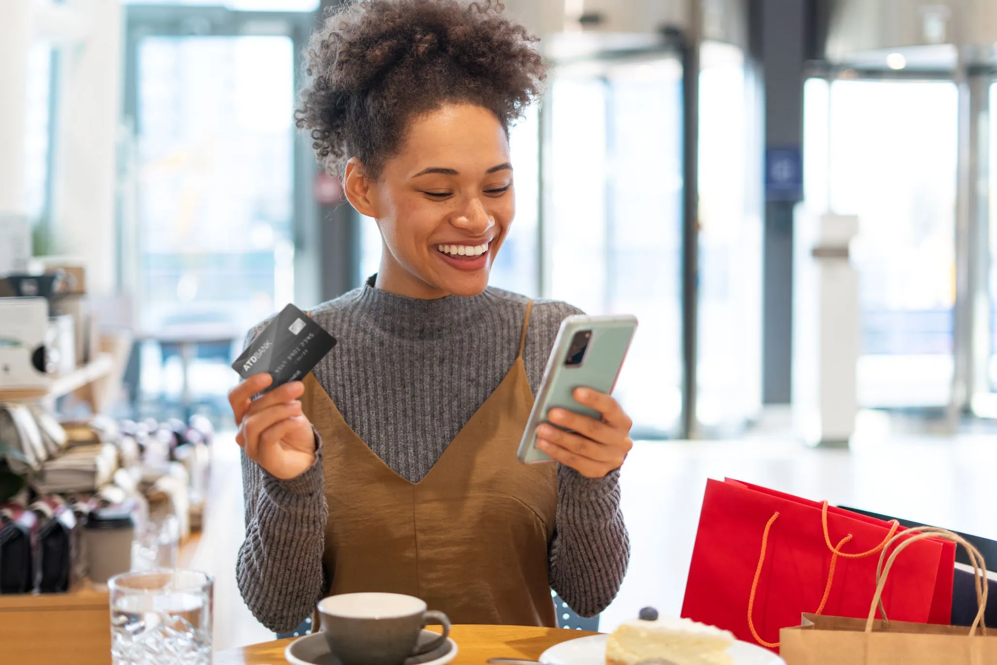 Mulher negra com suéter cinza está próxima a umas sacolas de compra segurando um celular com uma mão e na outra um cartão de crédito.