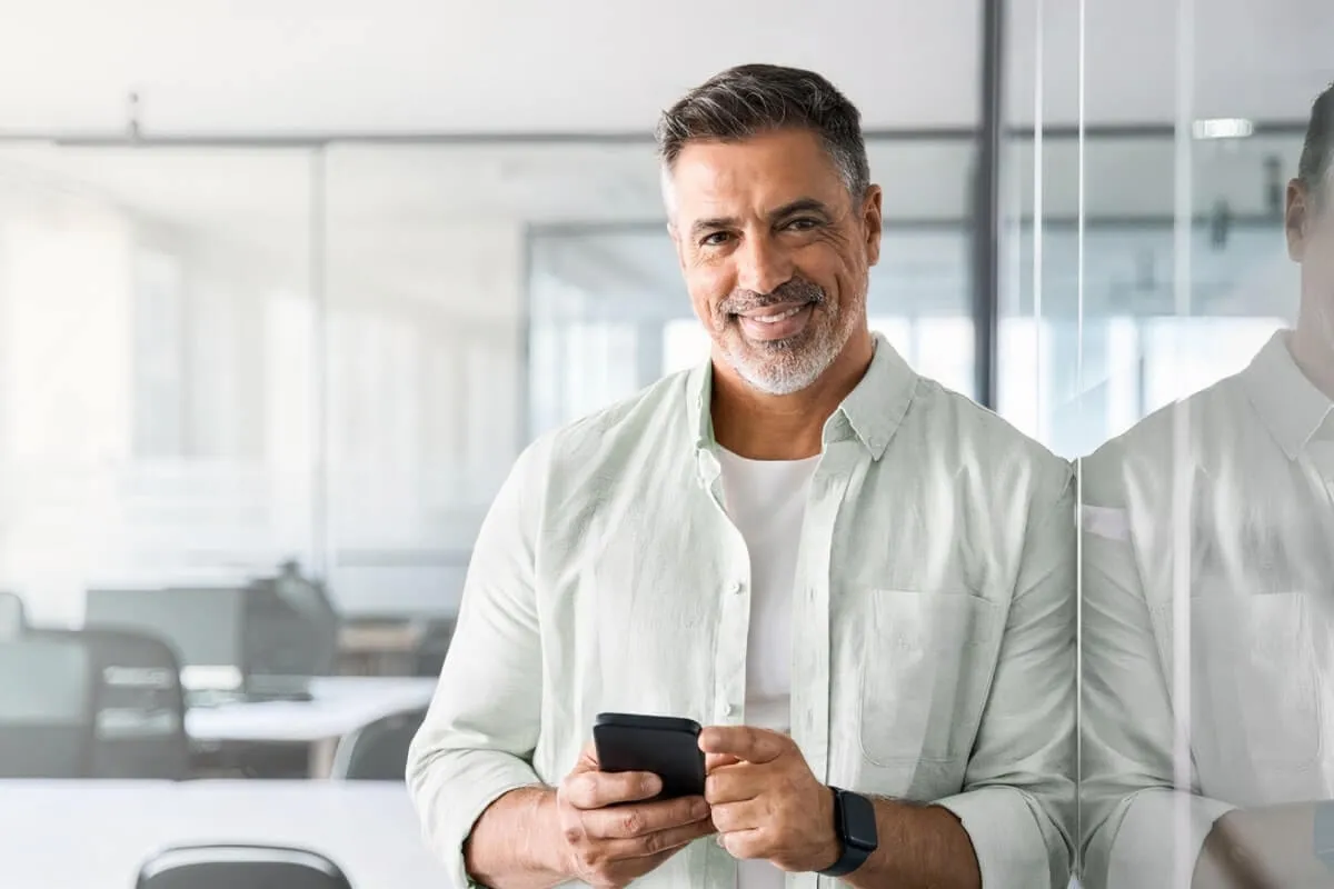 Homem sorrindo para câmera segurando um celular em seu escritório