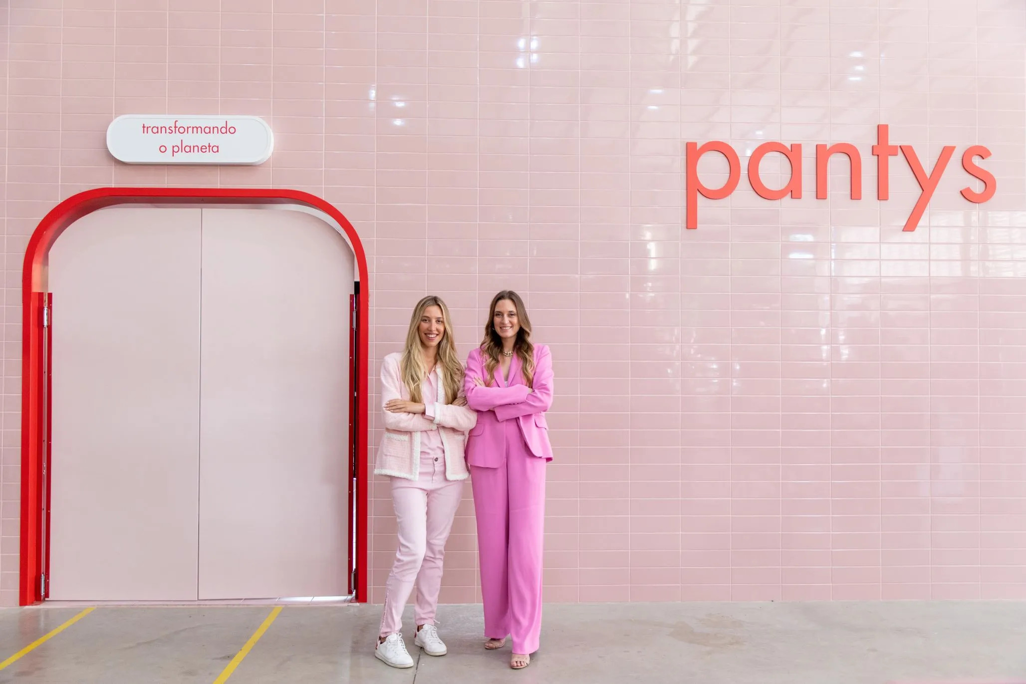 Duas mulheres brancas com o cabelo liso e loiro estão usando roupa cor-de-rosa, localizadas de pé em frente a uma parede cor-de-rosa claro escrito Pantys.