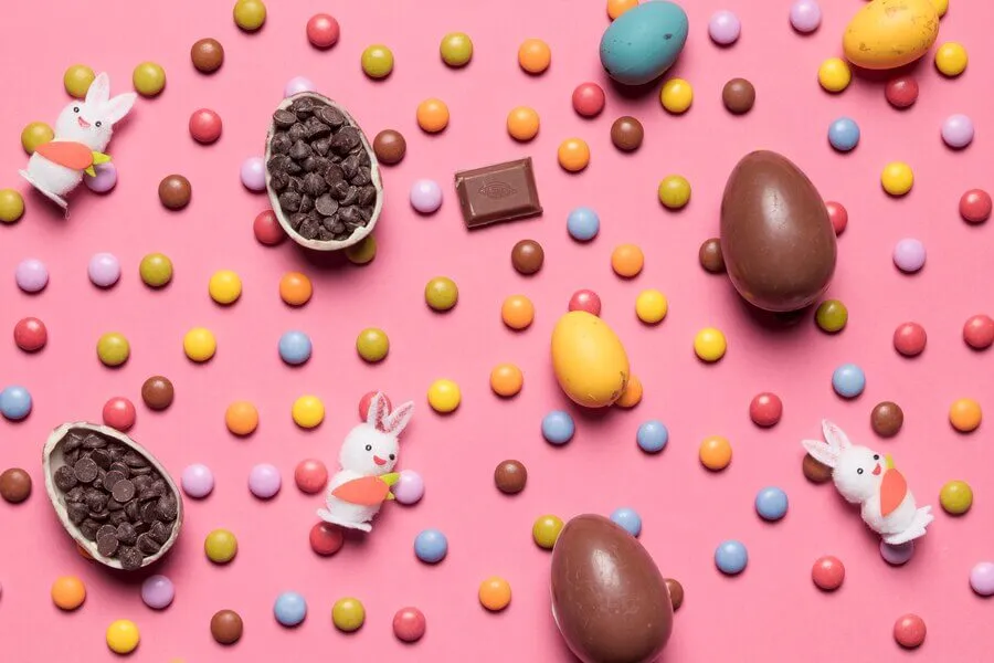 Ovos de chocolate, confeitos e pelúcias de coelho espalhadas por fundo rosa