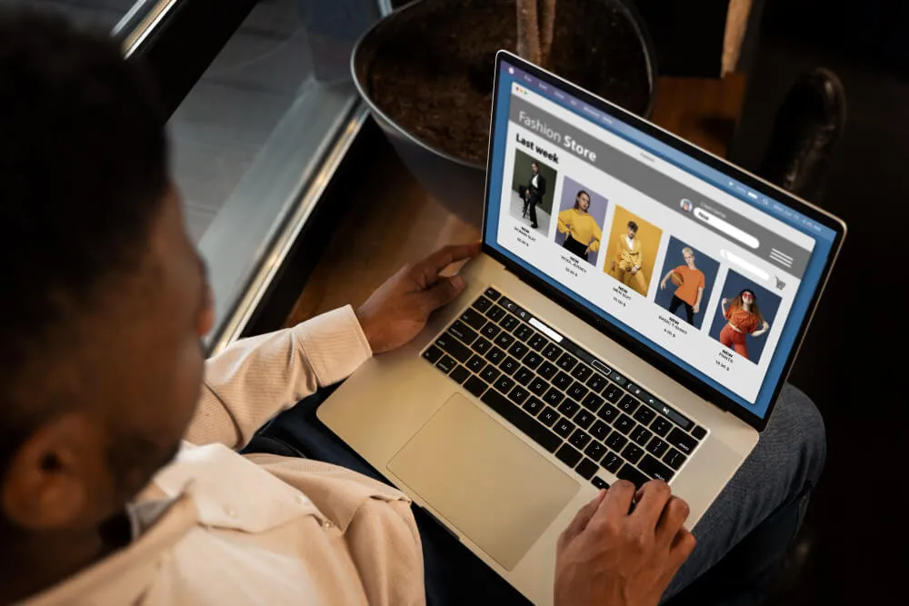 Homem negro sentado com computador no colo olhando site com roupas de moda 