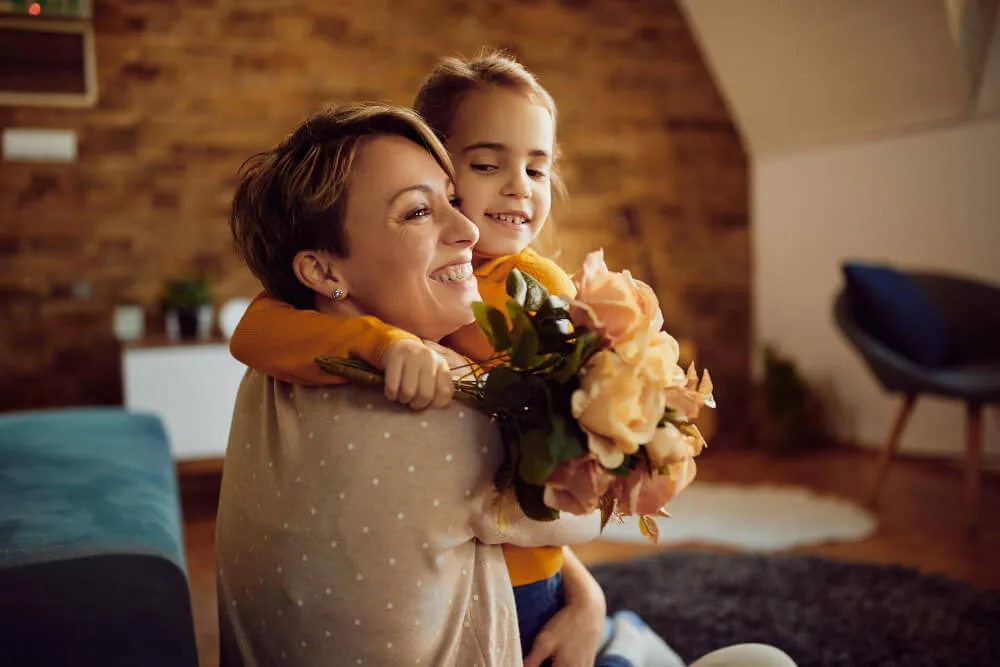 Mãe e filha, com flores em mãos, abraçadas pelo pescoço em comemoração ao dia das mães