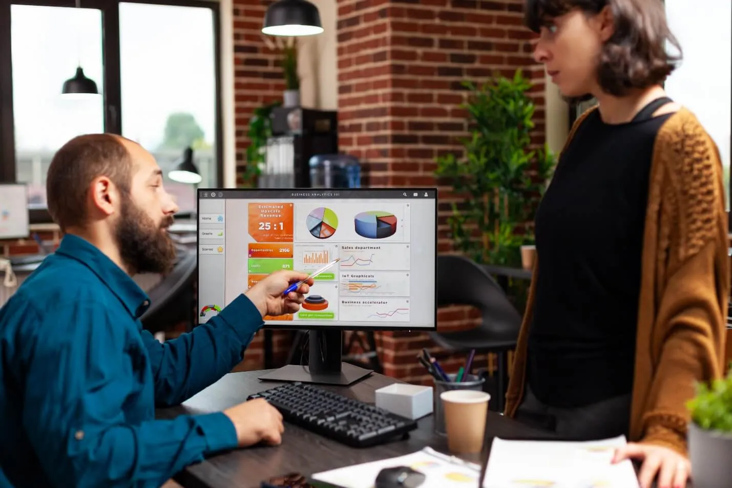 Homem branco e mulher branca conversando sobre gráficos e resultados mostrados na tela do computador
