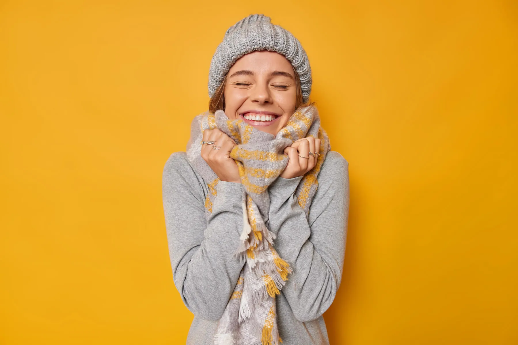 A woman smiles at a photo wearing winter clothes - hat, scarf and coat - on a yellow background