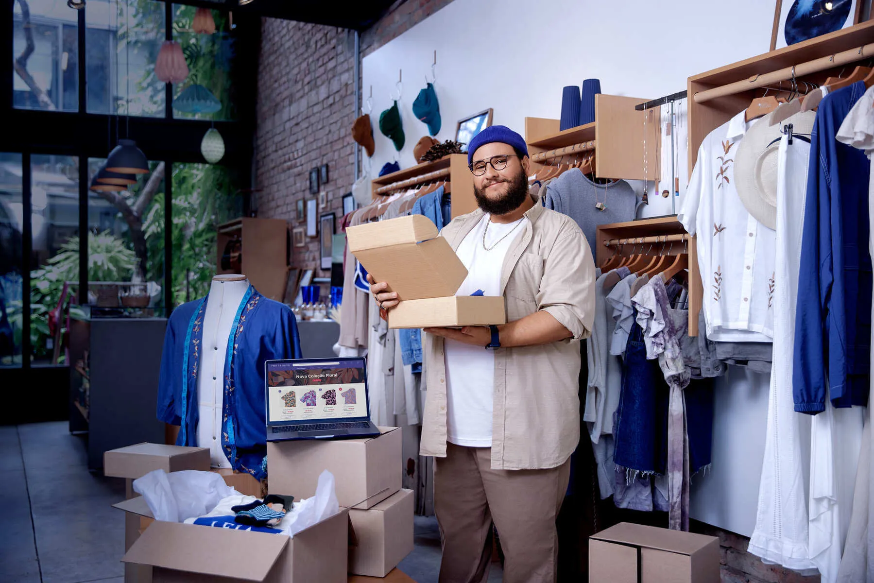 Empreendedor usando touca azul e camisa bege segurando caixa e prancheta em frente a arrara de roupas