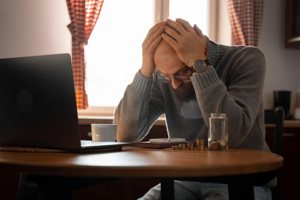 Homem frustrado, com as mãos na cabeça, de frente para o computador com pilhas de moedas em sua frente