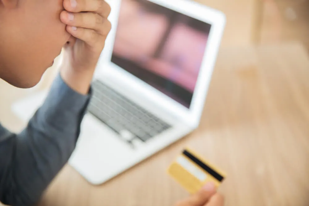 Consumidor com a mão nos olhos, frustrado com cartão de crédito em frente ao computador