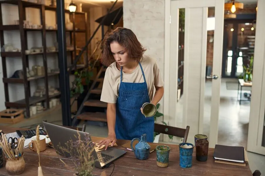 Mulher segurando pote de ceramica em mãos enquanto analisa pedidos em computador em estudio de produção