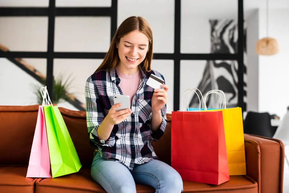 Mulher sentada no sofá fazendo compras pelo celular ao lado de sacolas