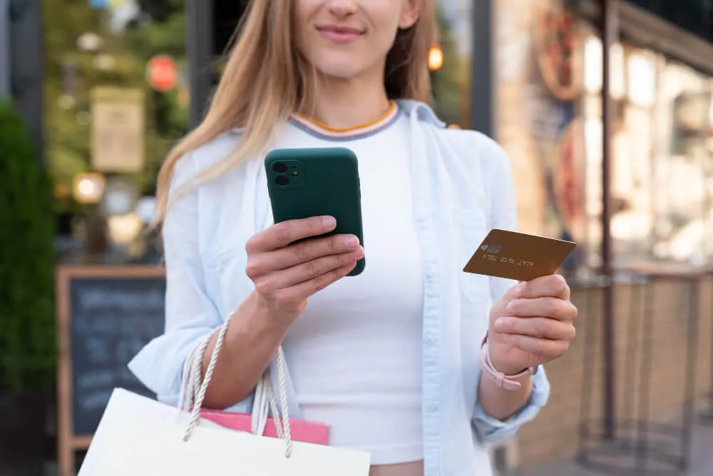 Mulher fazendo compras pelo celular com sacolas e cartão em mãos