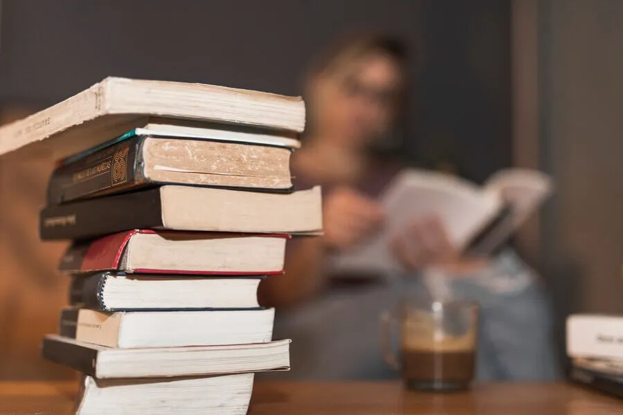 Pilha de livros ao lado de  mulher lendo com xícara de café em mesa de centro