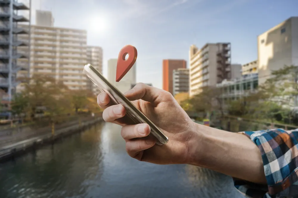 Pessoa segurando um smartphone próximo a um canal urbano, com um ícone de localização flutuando acima do aparelho.