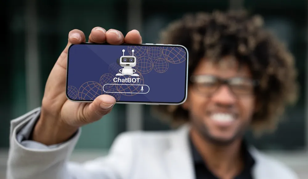 Homem sorridente segura um celular na horizontal, mostrando na tela um chatbot com um robozinho e o texto 'ChatBOT' sobre fundo azul.