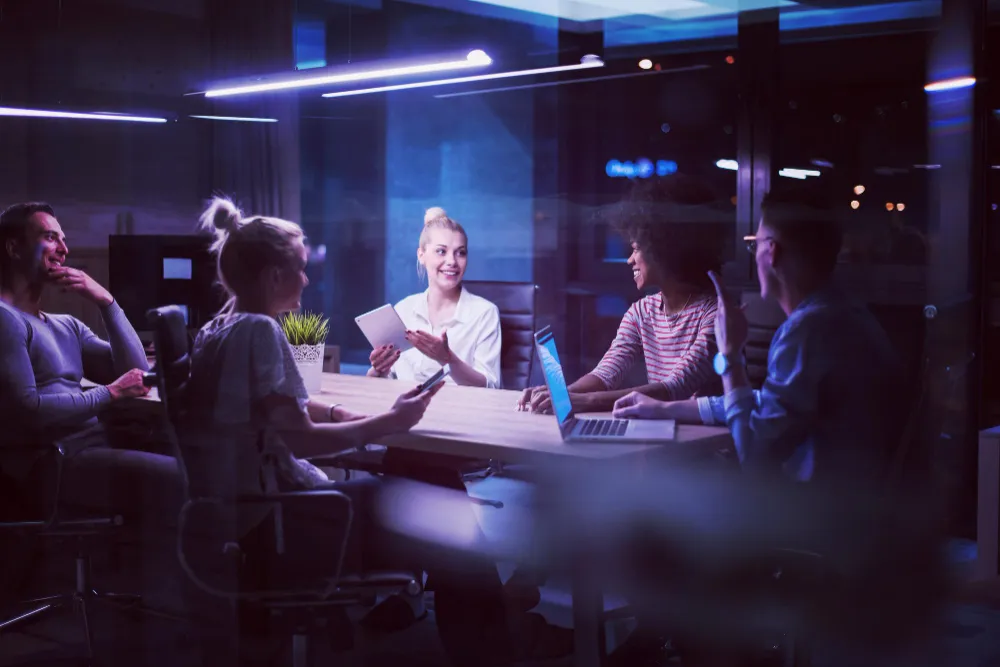 Grupo pessoas em reunião em uma sala de vidro, com clima descontraído, luz azul e uso de dispositivos eletrônicos.