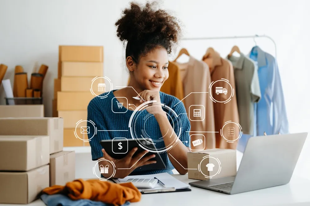 Mulher sorridente trabalhando em um pequeno negócio de e-commerce, cercada por caixas, roupas e dispositivos digitais, com ícones gráficos.