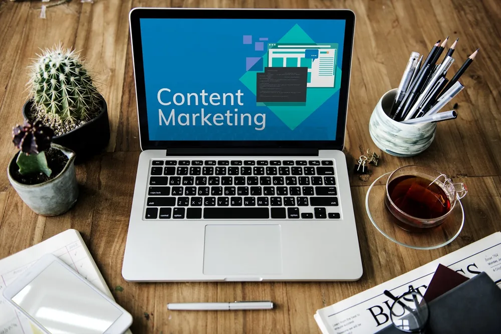 Notebook com a tela escrita "Content Marketing", sobre uma mesa com itens como chá, cactos, canetas e um jornal.