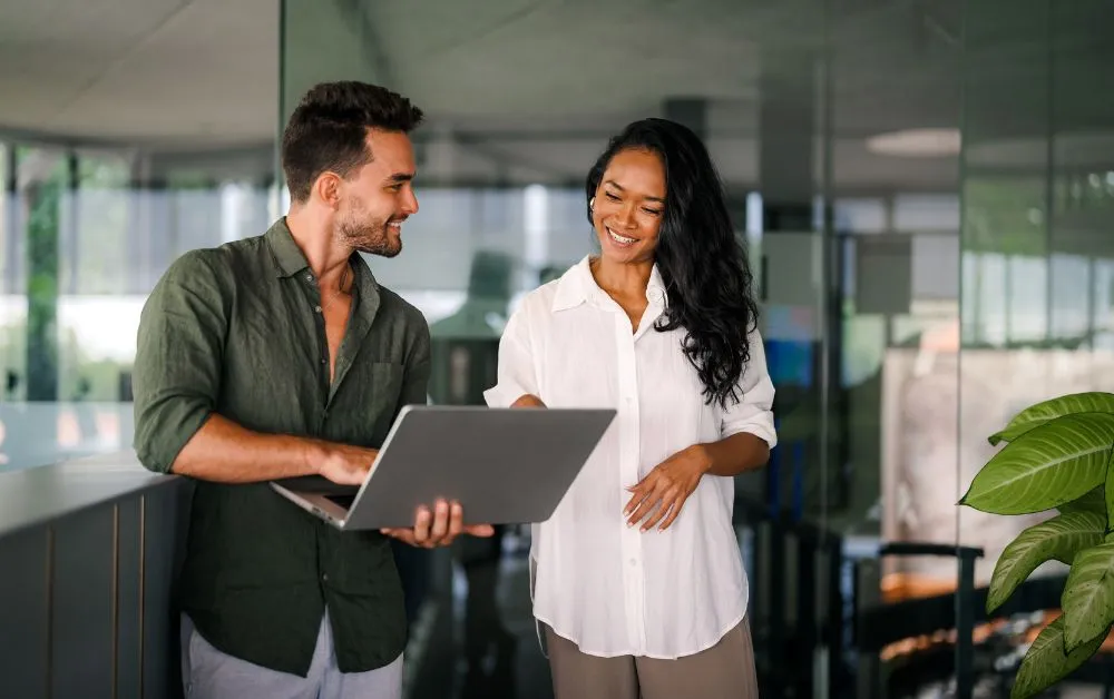Duas pessoas em um ambiente corporativo moderno, sorrindo enquanto olham para um notebook.