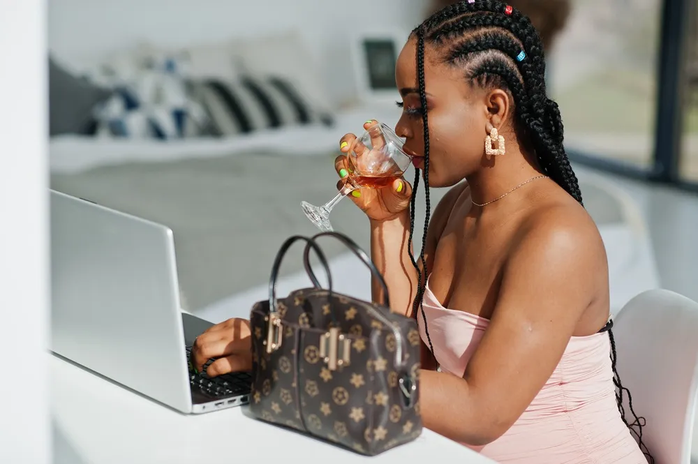 Mulher elegante trabalha no laptop enquanto bebe vinho, com bolsa de grife sobre a mesa.