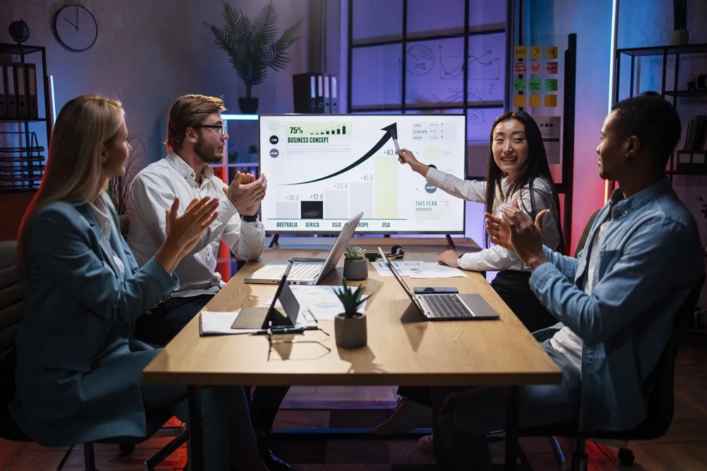 Pessoas em reunião aplaudindo enquanto uma mulher apresenta um gráfico de crescimento em um monitor.