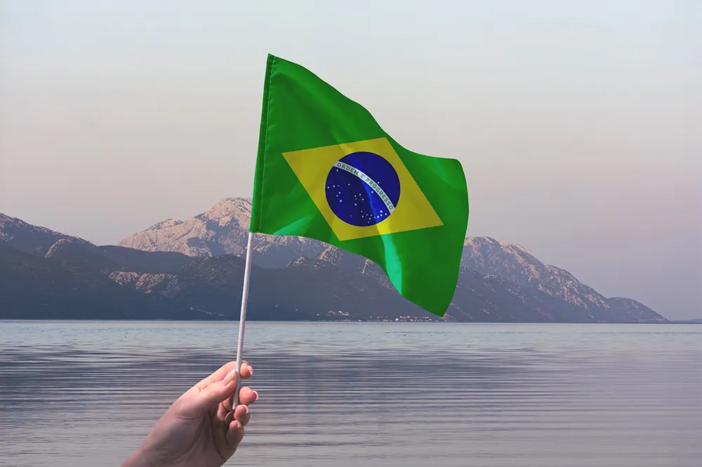 Bandeira do Brasil sendo segurada à mão diante de um lago com montanhas ao fundo.