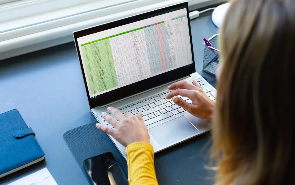Pessoa digitando em um notebook que exibe uma planilha com várias colunas coloridas.