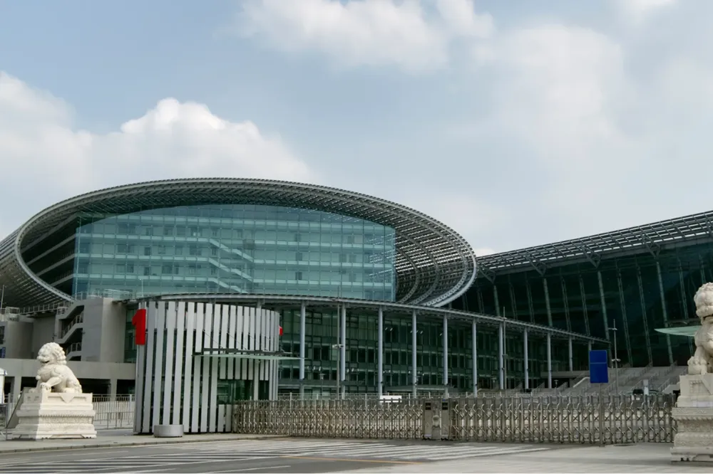 Fachada moderna da Canton Fair, com estrutura de vidro e aço, esculturas de leões e céu parcialmente nublado.