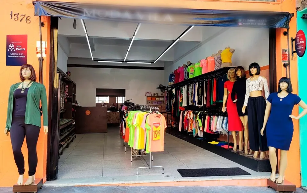 Fachada de loja de roupas com manequins femininos vestidos em diferentes estilos expostos na entrada e araras de roupas coloridas no interior.