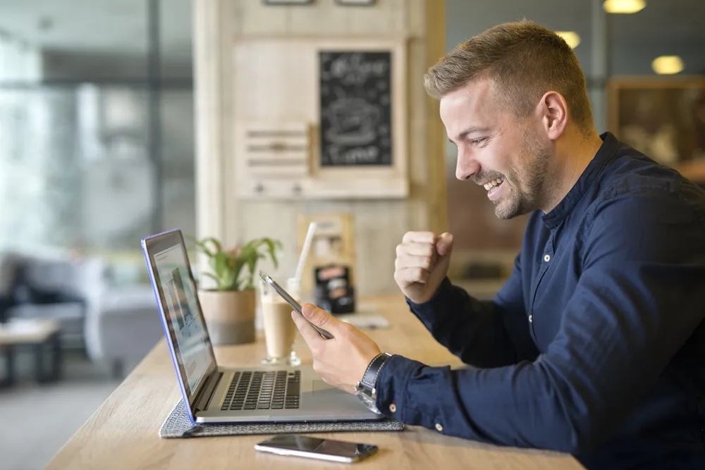 Homem sorridente comemorando enquanto olha para o celular, sentado em frente a um notebook em um café.