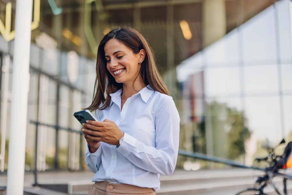 Mulher sorrindo enquanto usa o celular em frente a um prédio moderno.