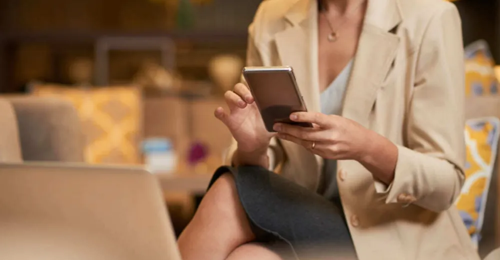 Pessoa sentada usando smartphone, com notebook aberto, em ambiente profissional.