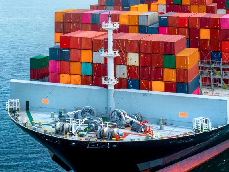 Parte frontal de um navio cargueiro navegando no mar, com diversos contêineres coloridos empilhados no convés.