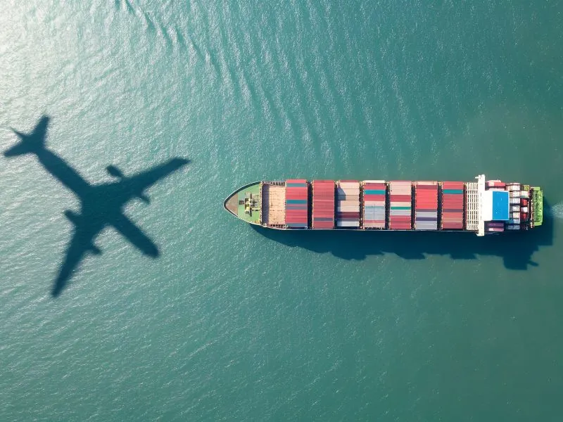 Vista aérea de navio cargueiro com contêineres no mar e sombra de avião na água.