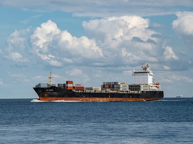 Navio cargueiro navegando em mar aberto, transportando contêineres empilhados no convés, sob céu com nuvens densas.