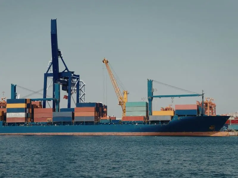 Vista lateral de um navio cargueiro azul atracado, com contêineres empilhados no convés e grandes guindastes portuários (um azul e outro amarelo) operando sob um céu claro.