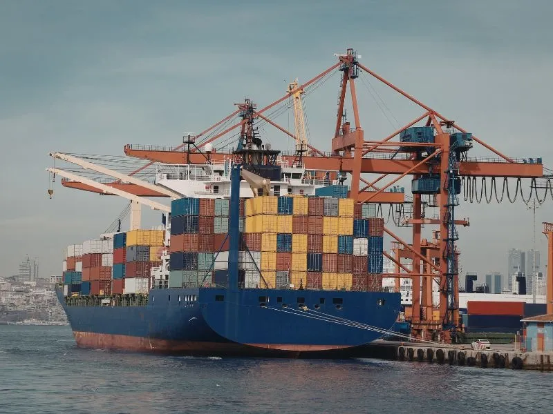 Vista da popa de um navio cargueiro azul atracado ao lado de grandes guindastes portuários laranjas, com destaque para as altas pilhas de contêineres coloridos e uma cidade ao fundo.