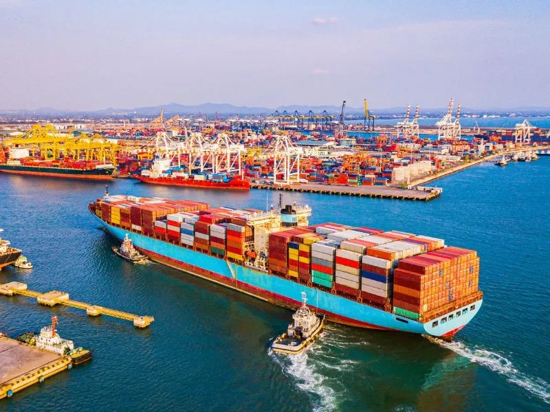 Vista panorâmica de um navio cargueiro azul sendo manobrado por rebocadores em um porto movimentado, com grandes guindastes amarelos e brancos e milhares de contêineres ao fundo.